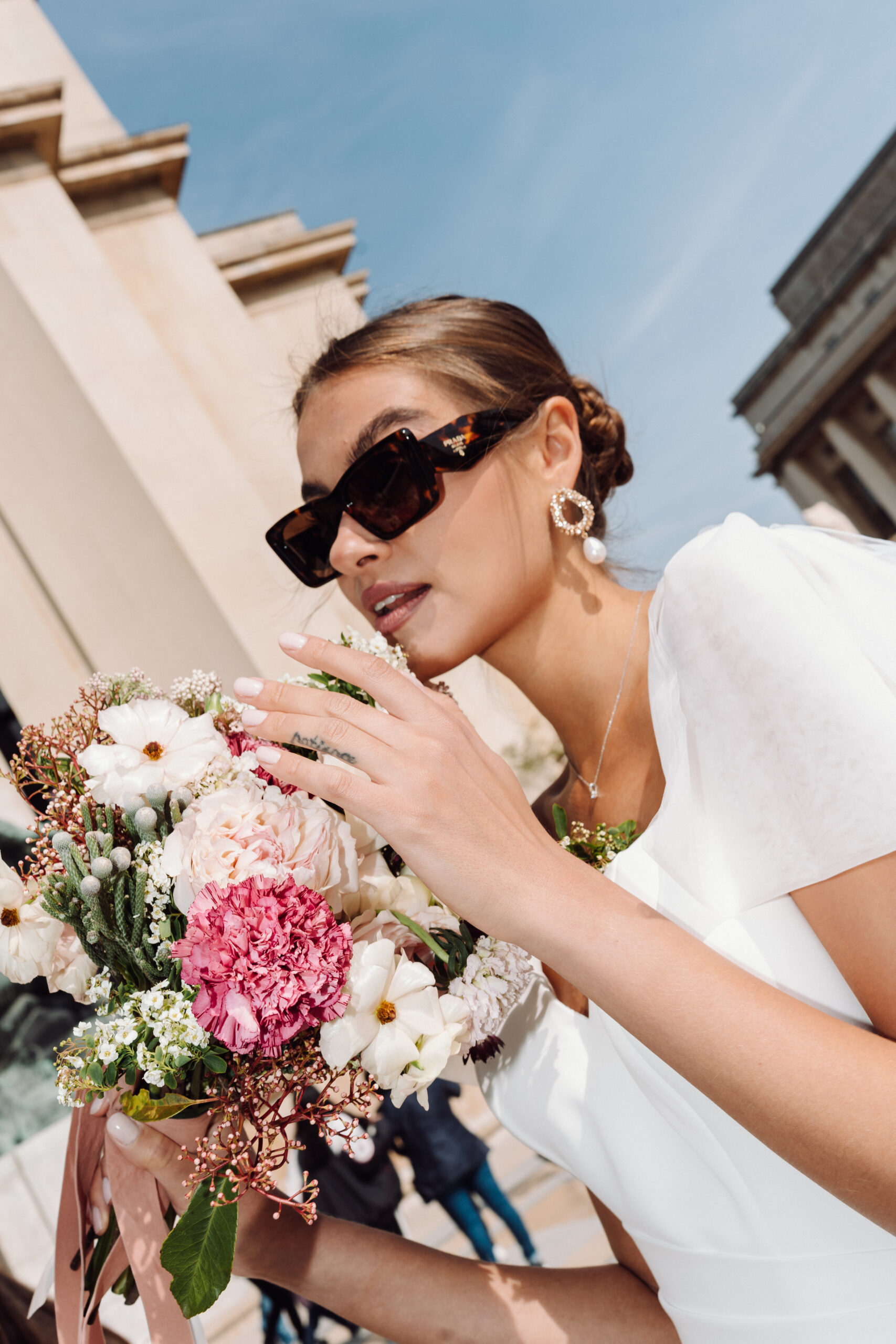 Frau in weißem Brautmode-Kleid mit Blumenstrauß und Sonnenbrille an einem sonnigen Tag im Freien - perfekte Inspiration vom Brautatelier bei Dornröschen.