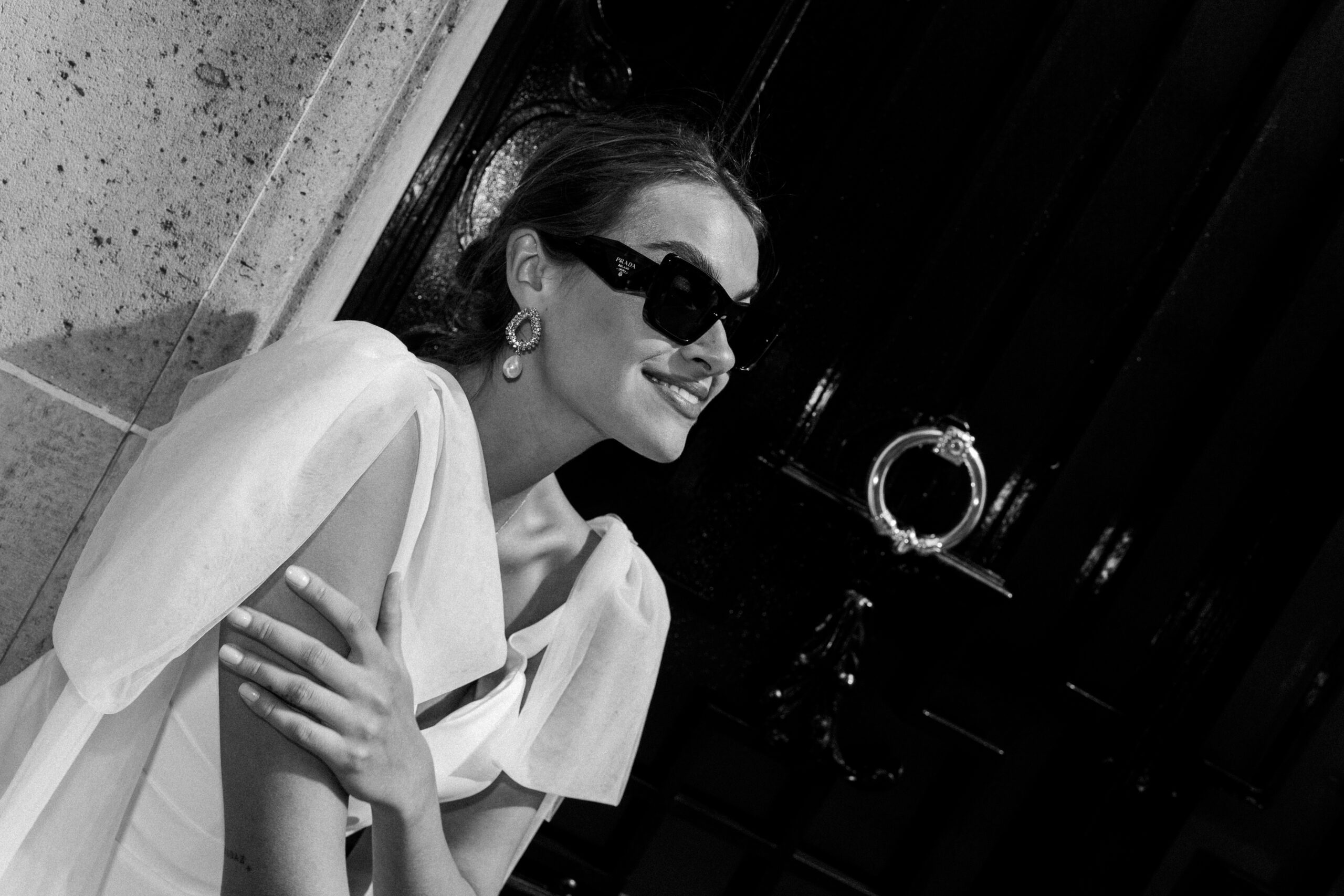 Frau mit Sonnenbrille und elegantem Kleid lehnt lächelnd an einer Wand vor einer dunklen Tür. Schwarz-Weiß-Foto, aufgenommen in der Nähe des Brautateliers bei Dornröschen in Nürnberg.