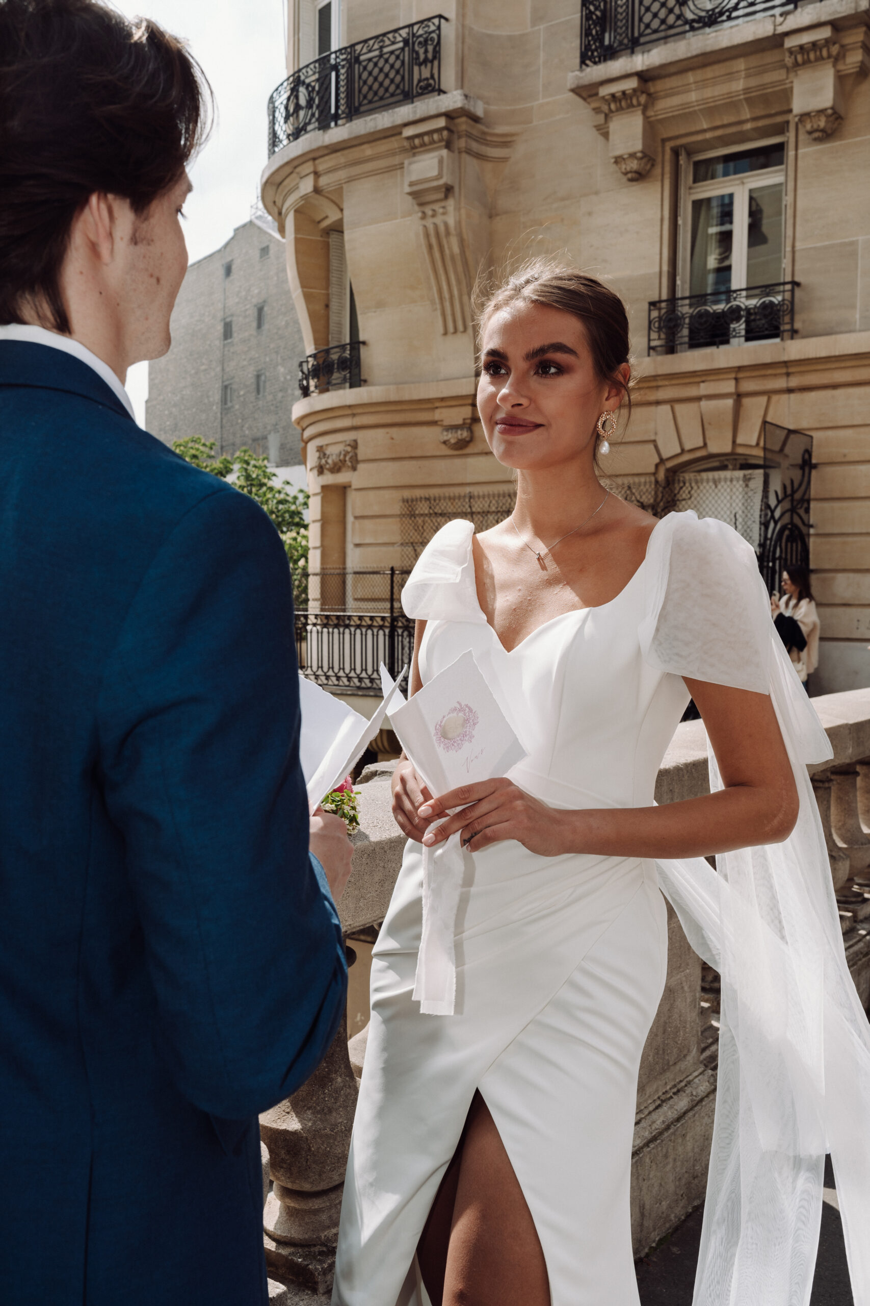 Braut und Bräutigam tauschen ihr Eheversprechen im Freien an einem steinernen Geländer aus, mit einem klassischen Gebäude im Hintergrund, das die elegante Brautmode bei Dornröschen in Nürnberg zeigt.
