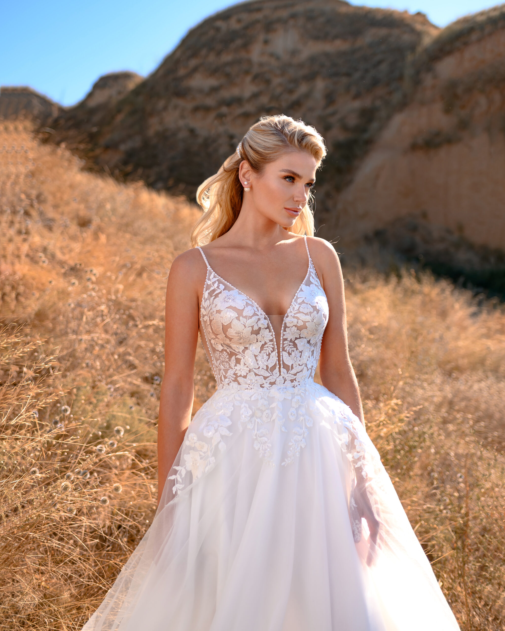Eine Frau in einem weißen Spitzenhochzeitskleid aus dem Brautatelier bei Dornröschen steht in einer trockenen, grasbewachsenen Landschaft mit felsigen Hügeln im Hintergrund.