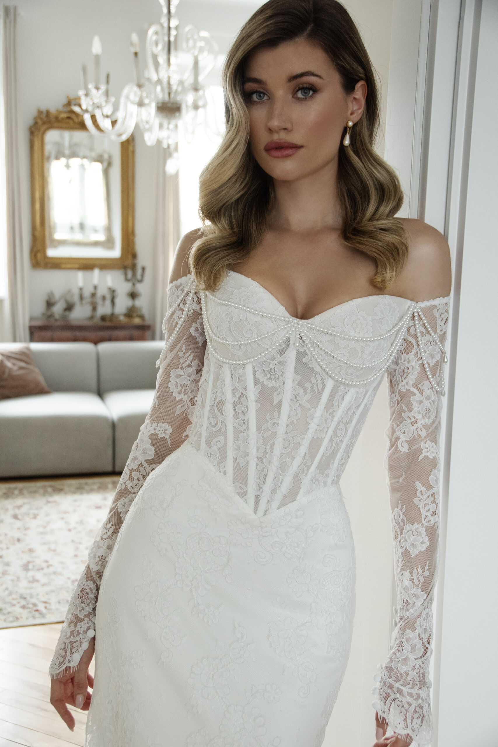 Frau in schulterfreiem Spitzenhochzeitskleid mit durchsichtigen Spitzenärmeln, stehend in einem hellen, eleganten Brautatelier in Nürnberg.