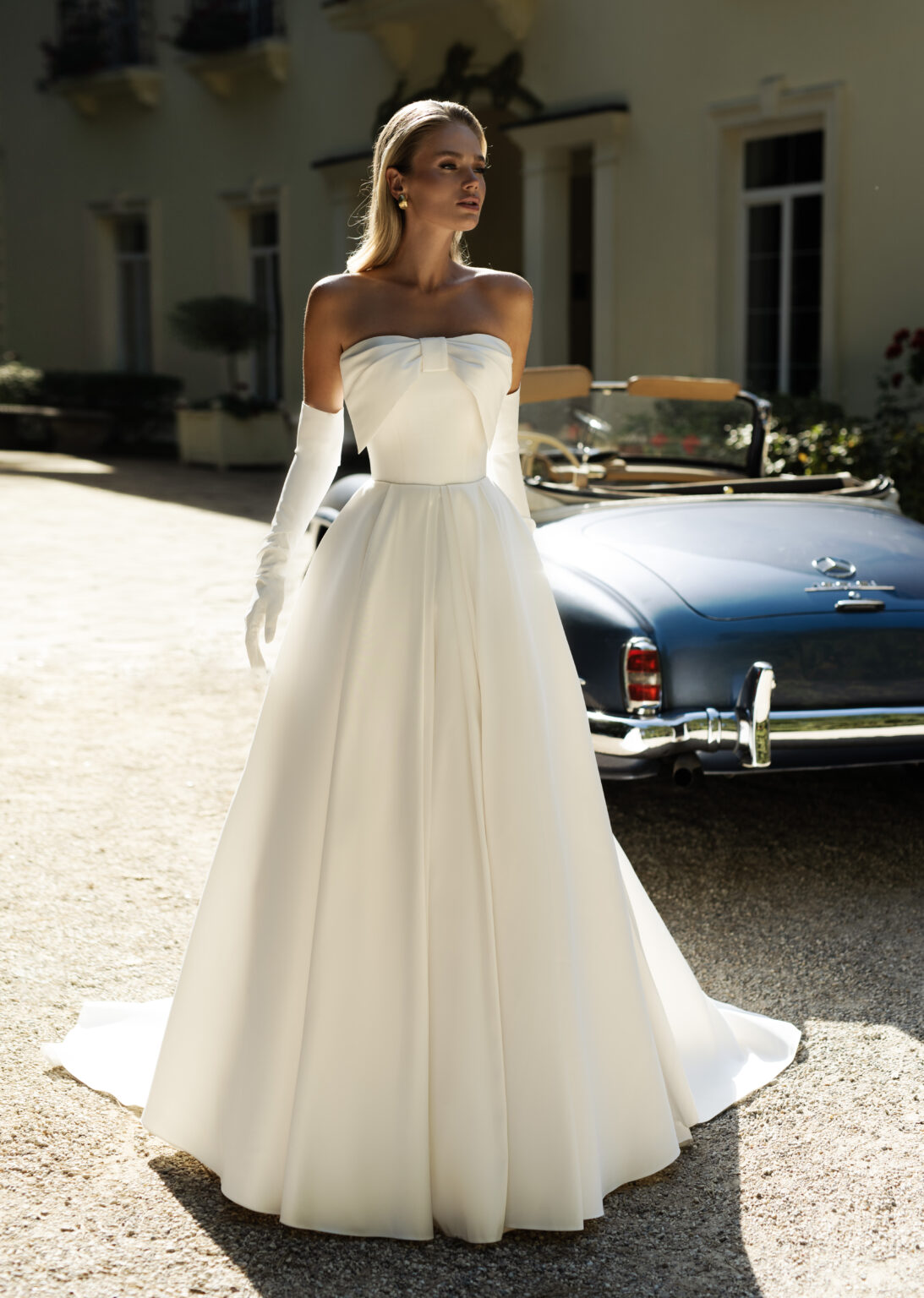 Eine Frau in eleganter Brautmode - einem weißen trägerlosen Kleid und Handschuhen - steht neben einem blauen Oldtimer-Cabrio und fängt zeitlosen Stil vor einem Gebäude in Nürnberg ein, in der Nähe eines renommierten Brautateliers.