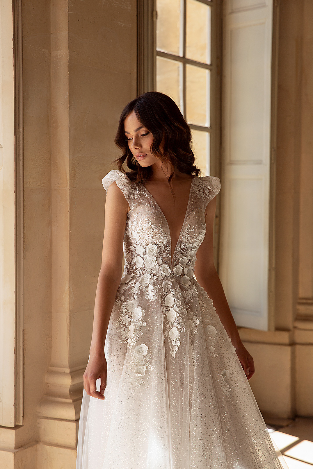 Eine Frau in einem Hochzeitskleid aus geblümter Spitze von Brautmode bei Dornröschen steht im weichen Sonnenlicht vor hohen Fenstern und Steinsäulen in Nürnberg.