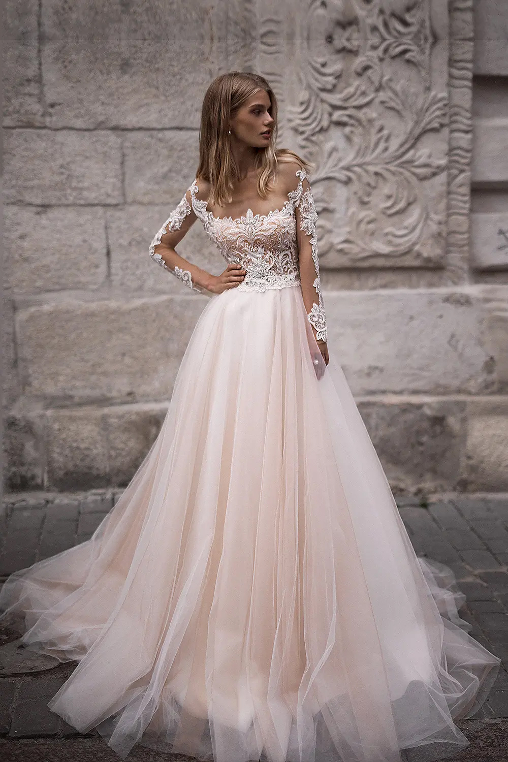 Eine Frau in einem schulterfreien Hochzeitskleid aus Spitze und Tüll steht vor einer strukturierten Steinwand und präsentiert elegante Brautmode aus einem renommierten Nürnberger Brautatelier.