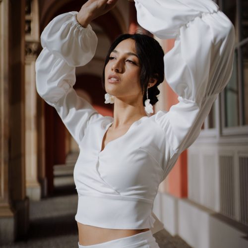 Eine Frau in weißem Outfit posiert im Freien mit erhobenen Armen und trägt markante Ohrringe unter einer bogenförmigen Architektur, die den eleganten Stil des Brautateliers bei Dornröschen in Nürnberg einfängt.
