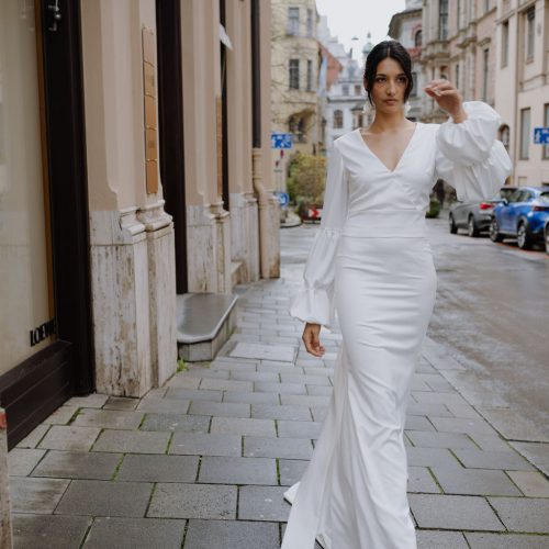 Eine Frau in einem weißen, langärmeligen Brautmode-Kleid von bei Dornröschen geht auf einem nassen Nürnberger Bürgersteig, der von historischen Gebäuden und parkenden Autos gesäumt ist.