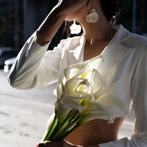 Eine Frau in weißem Gewand hält Calla-Lilien und bedeckt ihr Gesicht mit der Hand, während das Sonnenlicht Schatten wirft - ein heiterer Moment, inspiriert von der Brautmode in einem Nürnberger Brautatelier.