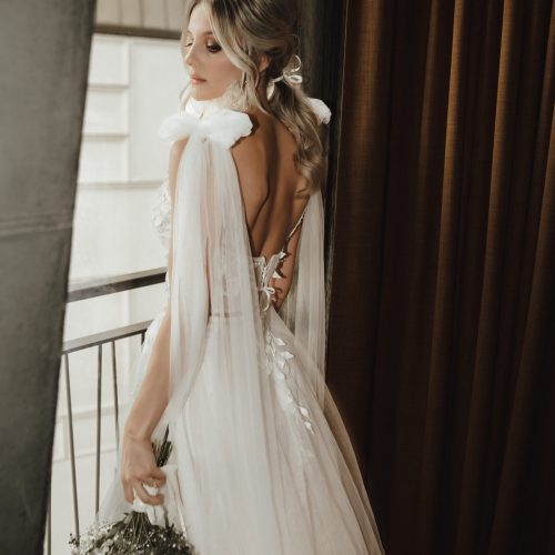Die Braut in einem fließenden weißen Kleid aus dem Brautatelier bei Dornröschen hält einen Blumenstrauß in der Hand und steht an einem Fenster mit durchsichtigen Vorhängen.