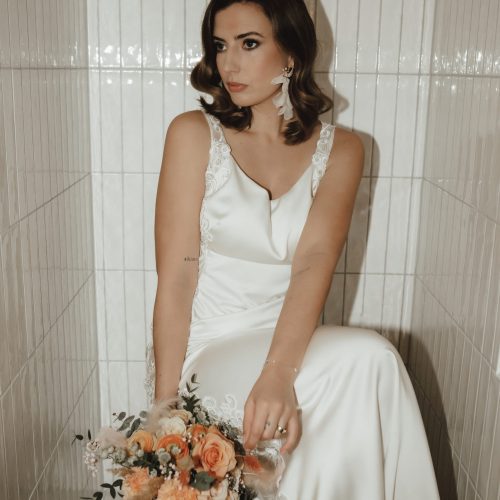 Eine Frau im weißen Satin-Hochzeitskleid von Brautmode sitzt in einem gekachelten Raum und hält einen Strauß orangefarbener Blumen - zeitlose Eleganz bei Dornröschen in Nürnberg.