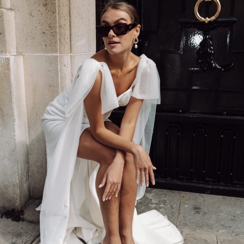 Eine Frau im weißen Kleid und mit Sonnenbrille, die an einer schwarzen Tür auf einer Steinstraße sitzt und stilvoll posiert - sie beschwört die Eleganz der Brautmode bei Dornröschen herauf, als würde sie aus einem Brautatelier auf die Straßen der Stadt treten.