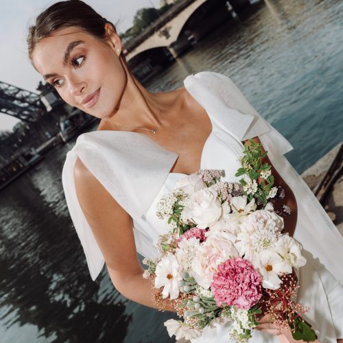 Die Braut in einem weißen Kleid mit einem Blumenstrauß in der Hand steht an einem Fluss mit einer Steinbrücke im Hintergrund in der Nähe von Nürnberg - ihr Kleid wurde vom Brautatelier bei Dornröschen entworfen.