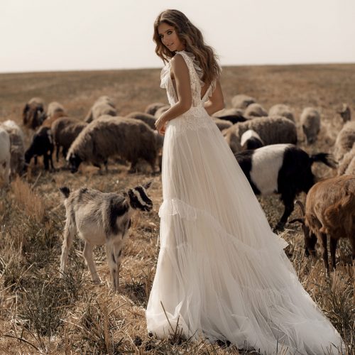 Eine Frau in einem wallenden weißen Kleid steht auf einer Wiese mit Schafen und einer Ziege um sie herum und erinnert an den märchenhaften Charme der Dornröschen Brautmode.