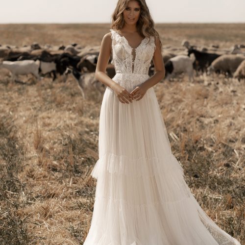Eine Frau in einem wallenden weißen Brautkleid aus Brautmode bei Dornröschen, Nürnberg, steht auf einem trockenen Feld mit einer Schafherde im Hintergrund.
