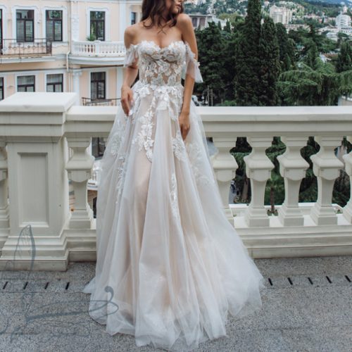 Eine Frau in einem schulterfreien Hochzeitskleid aus Spitze steht auf einer Terrasse mit Gebäuden und Grünflächen im Hintergrund und präsentiert elegante Brautmode aus einem Nürnberger Brautatelier.