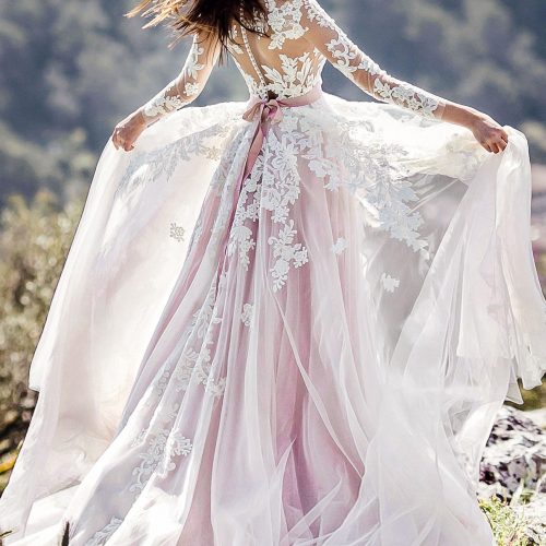 Eine Frau in einem fließenden, spitzenbesetzten Hochzeitskleid aus einem Nürnberger Brautatelier steht im Freien, hält ihren Rock in die Höhe und hat den Wind im Haar.