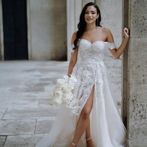 Braut in schulterfreiem Spitzenkleid mit Schlitz, weiße Rosen in der Hand, stehend an einer Steinsäule - elegante Brautmode wie im Brautatelier bei Dornröschen.