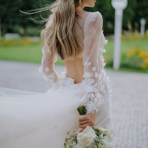 Eine Braut in einem durchsichtigen, floralen Kleid aus einem renommierten Nürnberger Brautatelier hält einen Strauß weißer Rosen im Freien, ihr Haar weht im Wind.