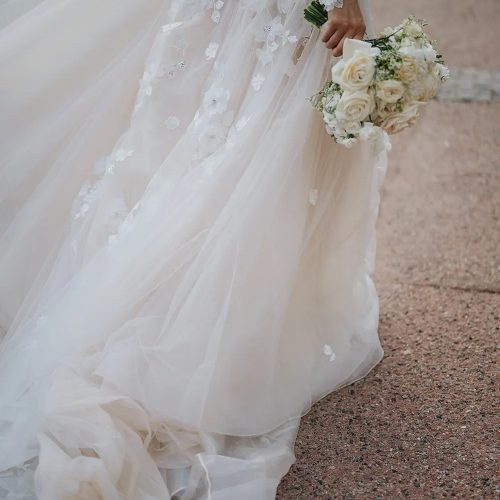 Die Braut in einem fließenden weißen Hochzeitskleid von Brautmode bei Dornröschen hält einen Strauß weißer Rosen, während sie einen Steinweg entlanggeht.
