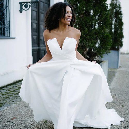 Eine lächelnde Frau in einem trägerlosen weißen Hochzeitskleid aus einem Brautatelier steht draußen in der Nähe von Grünflächen und einem weißen Gebäude in Nürnberg.