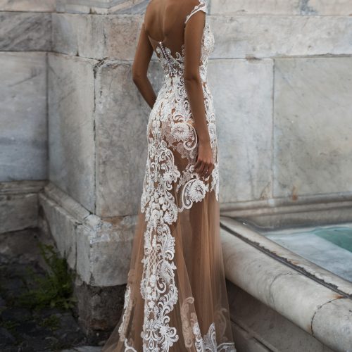 Eine Frau in einem eleganten, durchsichtigen Spitzenkleid steht an einer Steinmauer und blickt über ihre Schulter - sie fängt die zeitlose Schönheit der Brautmode in einem Brautatelier in Nürnberg ein.