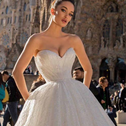 Eine Frau in einem trägerlosen weißen Hochzeitskleid posiert im Freien vor einem historischen, verschnörkelten Steingebäude, das elegante Brautmode bei Dornröschen zeigt.